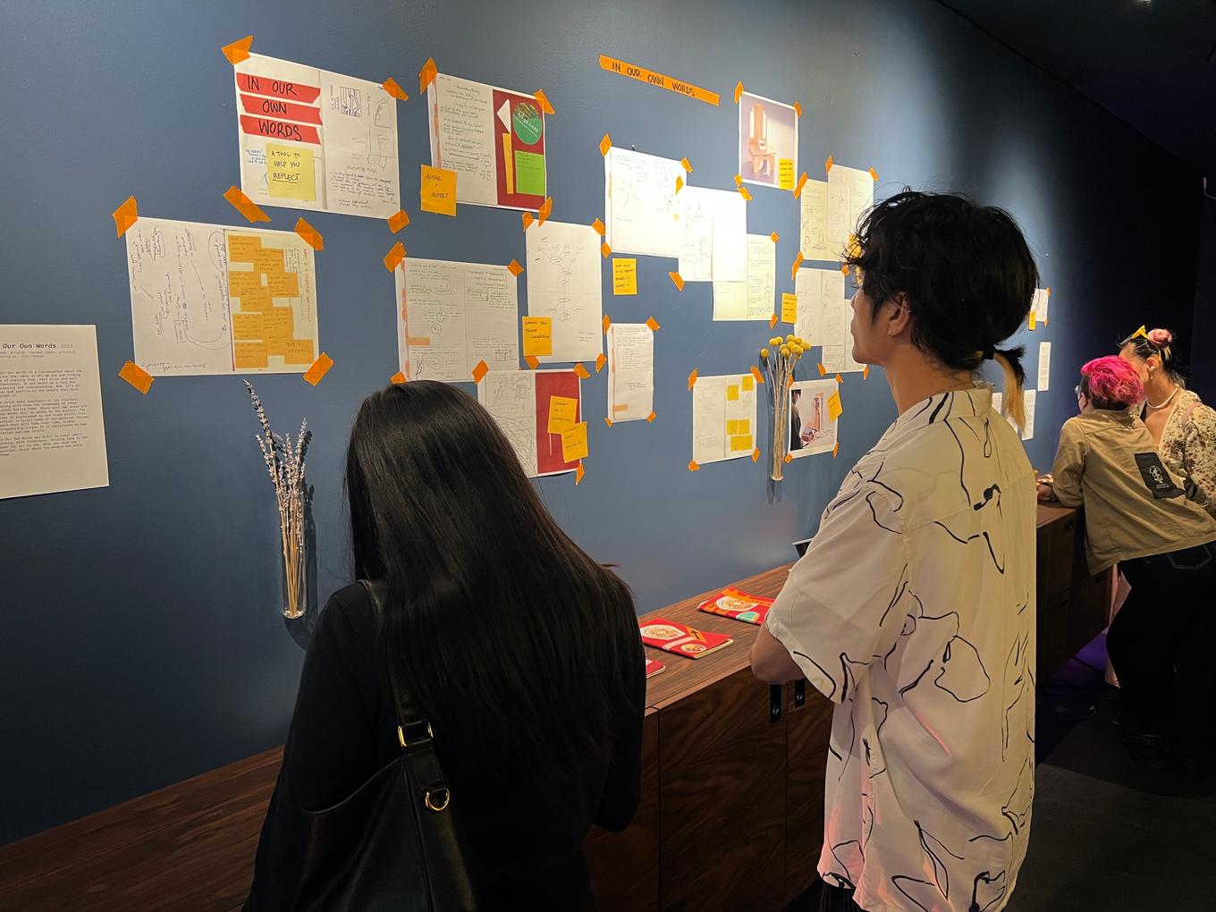 Two figures standing in front of a wall that is covered in many sheets of paper, taped up. The paper appears to show sketches.