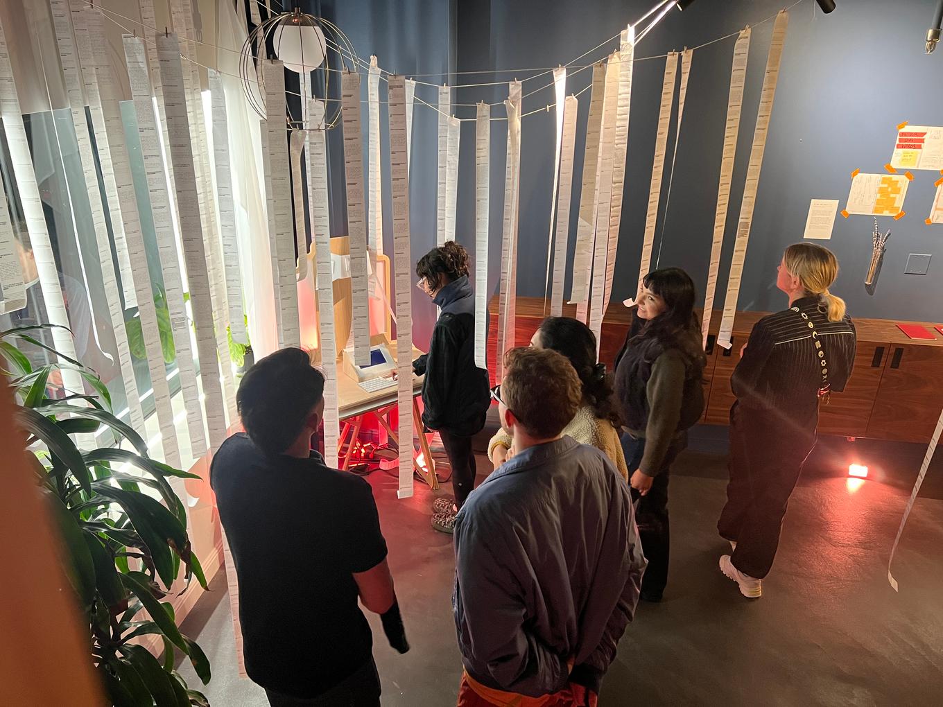 Many figures standing, surrounded by draped receipt paper. One person is standing at an altar, hand on the keyboard.